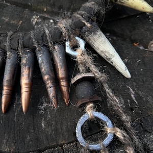 Op de afbeelding: Een ketting gemaakt van bruin touw, metalen ringen en patroonhulzen. De ketting heeft ook een kleine, donkerbruine houten kraal en een witte, tandenvormige hanger.