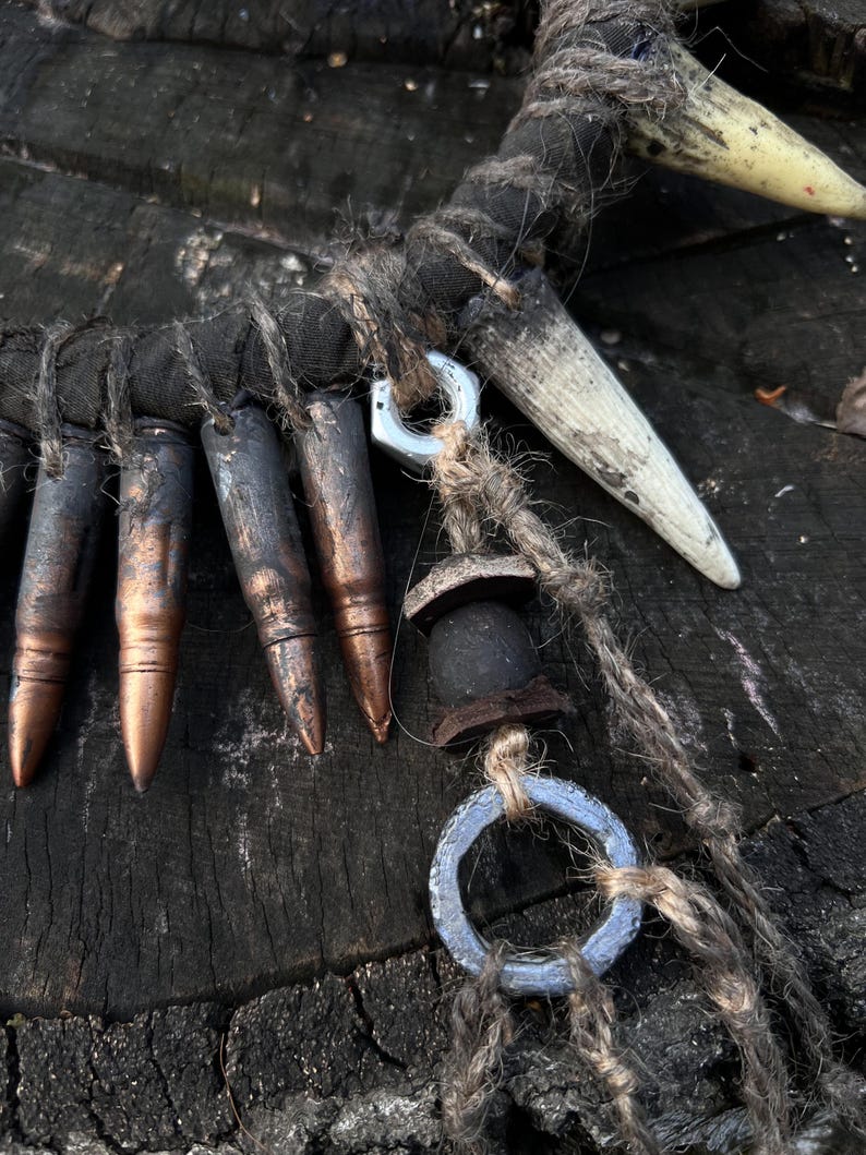 Op de afbeelding: Een ketting gemaakt van bruin touw, metalen ringen en patroonhulzen. De ketting heeft ook een kleine, donkerbruine houten kraal en een witte, tandenvormige hanger.