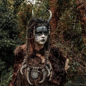 May include: A woman wearing a brown fur coat with a silver necklace and horns. She has black and white face paint and long black hair.