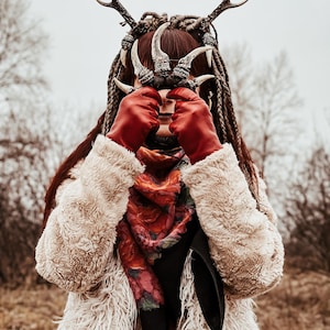 Peut inclure: Une personne portant un manteau en fausse fourrure blanc avec une écharpe rouge et un couvre-chef en bois de cerf décoratif. La personne se couvre le visage avec des gants rouges.