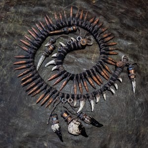 Op de afbeelding: Een handgemaakte spiraalvormige ketting met koperkleurige hulzen, witte tandachtige elementen en zwarte stof. De ketting heeft een ruig uiterlijk, met metalen en benen accenten.