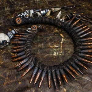 Op de afbeelding: Een ketting gemaakt van kogelhulzen, gewikkeld in donkerbruin touw, met een schedelhanger en andere metalen accenten.