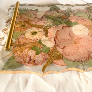 May include: A clear resin tray with a gold metal handle. The tray is filled with pressed flowers and leaves in shades of pink, green, and gold.