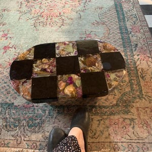 Pode incluir: Mesa de centro oval com um design de xadrez. A superfície da mesa apresenta quadrados pretos alternados e quadrados preenchidos com flores secas e resina. A mesa está sobre um tapete estampado.