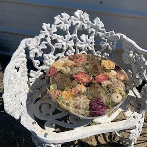 Pode incluir: Uma peça redonda de resina transparente com flores secas e ouriços-do-mar. As flores são de várias cores, incluindo rosa, amarelo e creme. A peça de resina está sobre um banco de metal branco ornamentado.