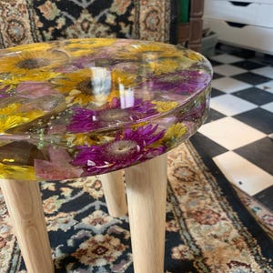 Pode incluir: Uma mesa lateral redonda com um tampo de resina transparente com flores amarelas e roxas embutidas. A mesa tem três pernas de madeira clara. A mesa tem cerca de 46 cm de diâmetro.