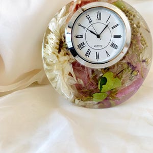 May include: A round, clear clock with a silver face and Roman numeral markings. The clock is embedded with dried flowers and greenery.