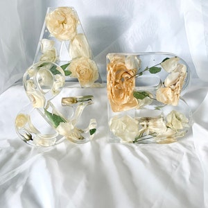 May include: Two clear resin letters, "A" and "&", filled with dried white and cream flowers and greenery.