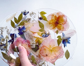 May include: A clear resin coaster with pressed flowers and leaves. The flowers are pink, yellow, and blue. The leaves are green.