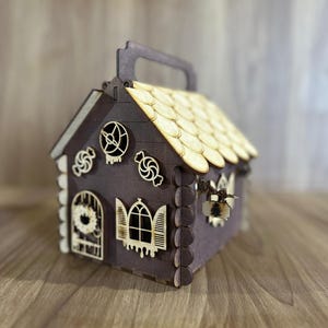 Puede incluir: Una caja de madera con forma de casa de jengibre con asa. La casa es de color marrón oscuro con un tejado marrón claro y recortes decorativos de caramelos, una puerta y ventanas. El tejado está hecho de pequeñas tejas de madera superpuestas.