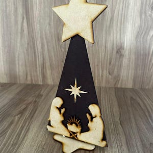 Puede incluir: Adorno de árbol de Navidad de madera con una estrella en la parte superior, una base triangular marrón oscuro y una escena de la Natividad. Las figuras están cortadas de madera de color claro, con una estrella en el centro. La decoración está colocada sobre una superficie de madera.