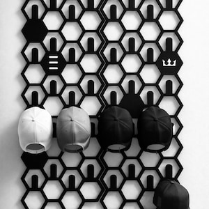May include: Black hexagonal wall-mounted hat rack with several baseball caps. The rack is made of black metal and features a honeycomb design. The caps are white, grey, and black. The rack has white logos.