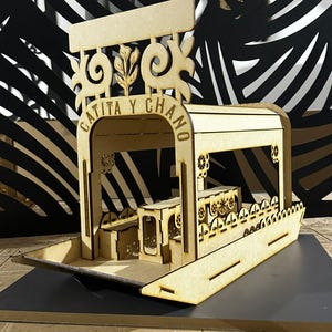 Puede incluir: Maqueta de madera de un barco con el texto "CATITA Y CHANO" en el techo. El barco tiene cortes decorativos y un interior detallado, incluyendo una pequeña mesa. El modelo es de madera marrón claro y está sobre una superficie oscura.