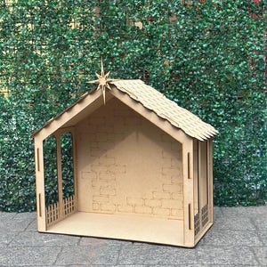 May include: A wooden nativity scene, featuring a small stable with a pitched roof and a star on top. The structure has a brick-like pattern on the walls and a small fence in front. The overall color is a light brown.
