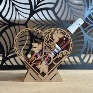 May include: A wooden heart-shaped wine bottle holder with laser-cut grape vine details. The holder is made of light brown wood and holds a bottle of wine. The background features a decorative black metal leaf design.