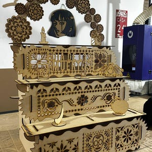 Puede incluir: Una estantería de madera cortada con láser de varios niveles con intrincados diseños del Día de Muertos. La estantería presenta recortes decorativos de flores, calaveras y el texto "DIA DE LOS MUERTOS". Los recortes de madera adicionales incluyen una vela, una cara de mujer y una manzana.