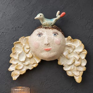 Op de afbeelding: Een keramieken wandsculptuur met een rond gezicht, roze wangen, blauwe ogen en een koraalkleurige mond. De figuur heeft bruin haar met een kleine blauwe en koraalkleurige vogel erbovenop, en grote, gestructureerde, crèmekleurige vleugels.