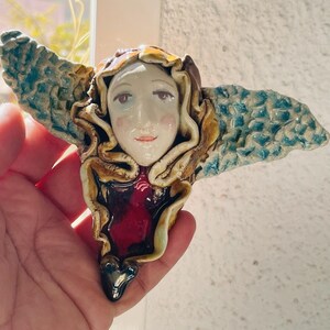 May include: A ceramic angel figurine with a serene face, flowing hair, and turquoise wings. The angel's face is painted with delicate features, and the wings have a textured, patterned design. The figurine has a red heart-shaped detail and a black heart at the bottom.