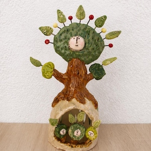 Könnte beinhalten: Eine Keramikskulptur eines Baumes mit einem Gesicht auf dem Stamm und drei kleineren Gesichtern in einer hohlen Basis. Der Baum hat grüne Blätter, rote und gelbe Beeren an Drahtstielen und einen strukturierten braunen Stamm. Die Basis ist cremefarben.