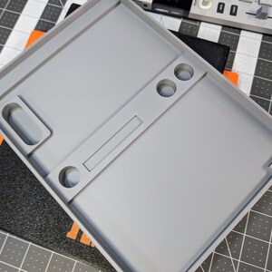 May include: A light gray, rectangular tray with various compartments and recessed areas. The tray has a raised border and includes circular and rectangular cutouts. The design suggests a tool or organizational tray, possibly for small parts or components.