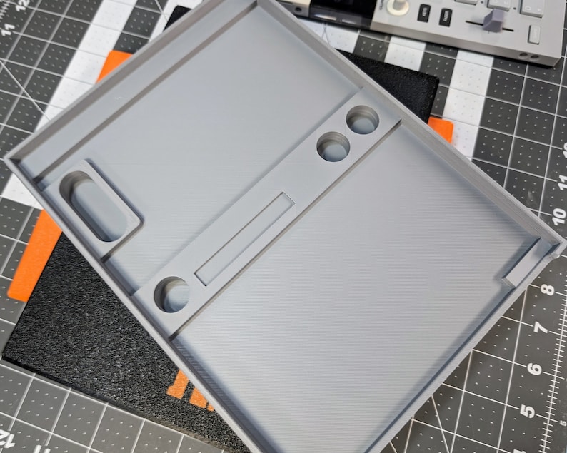 May include: A light gray, rectangular tray with various compartments and recessed areas. The tray has a raised border and includes circular and rectangular cutouts. The design suggests a tool or organizational tray, possibly for small parts or components.
