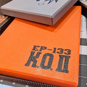 May include: An orange rectangular pad with the text "EP-133 K.O.II" in black. A gray pad and a black pad are stacked on top. The pads appear to be made of foam or a similar material. The background includes a cutting mat.