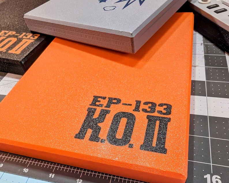 May include: An orange rectangular pad with the text "EP-133 K.O.II" in black. A gray pad and a black pad are stacked on top. The pads appear to be made of foam or a similar material. The background includes a cutting mat.