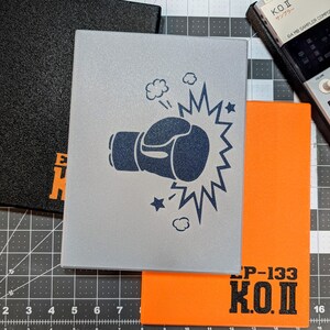 May include: Three rectangular notebooks in black, silver, and orange. The silver notebook features a boxing glove graphic with a burst. The orange notebook has "LP-133 K.O.I" printed on it. A black notebook with orange text is also visible. A sampler is in the background.
