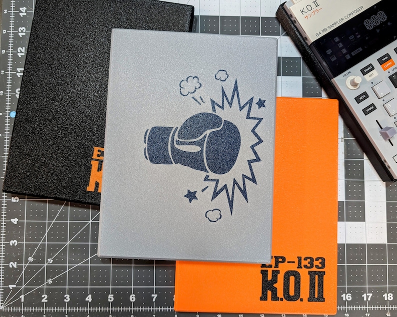 May include: Three rectangular notebooks in black, silver, and orange. The silver notebook features a boxing glove graphic with a burst. The orange notebook has "LP-133 K.O.I" printed on it. A black notebook with orange text is also visible. A sampler is in the background.