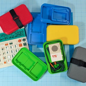 Puede incluir: Un surtido de contenedores de plástico de colores y dispositivos electrónicos. Un contenedor rojo con una correa negra se encuentra encima de un dispositivo blanco y verde con la etiqueta "RIDDIM". Otros contenedores son azules, amarillos, verdes y grises.