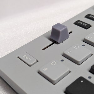 May include: Close-up of a gray audio mixer control panel. The panel features various buttons and sliders, including a prominent gray slider knob. Buttons are labeled "SHIFT", "FADER", "KEYS", and "TUNE".