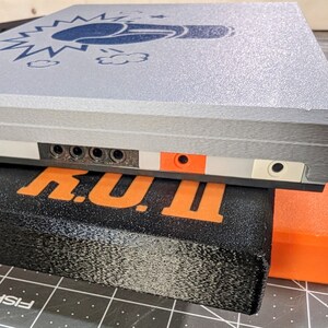 May include: A gray rectangular device with a blue graphic of a fist and lightning bolts. The device has multiple ports and is resting on a black rectangular object with the word "TINY" in orange. An orange rectangular object is also visible.