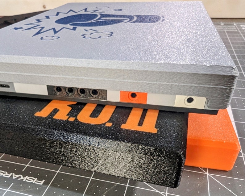 May include: A gray rectangular device with a blue graphic of a fist and lightning bolts. The device has multiple ports and is resting on a black rectangular object with the word "TINY" in orange. An orange rectangular object is also visible.