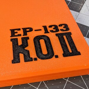 May include: Close-up of an orange rectangular object with the text "EP-133 K.O. II" printed in black. The object has a textured surface and appears to be a piece of equipment or a component.