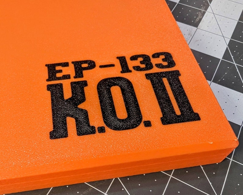 May include: Close-up of an orange rectangular object with the text "EP-133 K.O. II" printed in black. The object has a textured surface and appears to be a piece of equipment or a component.