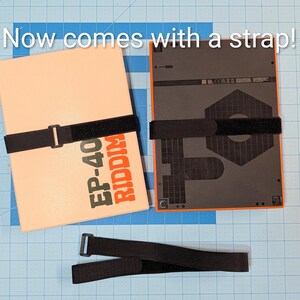 May include: Two rectangular electronic devices with black straps, one cream-colored with orange text reading "EP-40 RIDDIM", the other black with an orange border. A separate black strap is also visible. Text at the top reads "Now comes with a strap!"