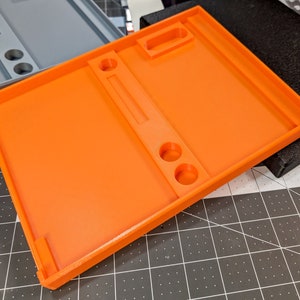 May include: Bright orange 3D-printed tray with a rectangular shape. The tray features a raised border, a central divider, two circular holes, and a small rectangular cutout. A gray tray is visible in the background.