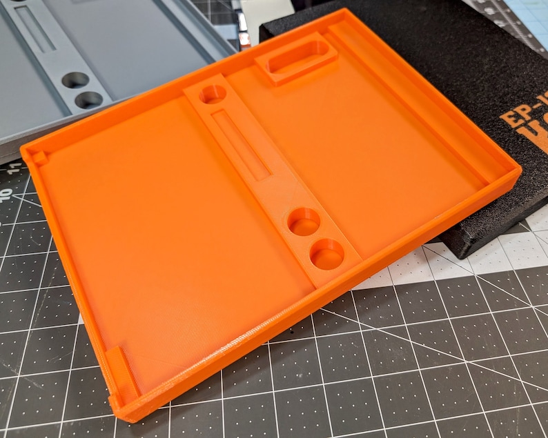 May include: Bright orange 3D-printed tray with a rectangular shape. The tray features a raised border, a central divider, two circular holes, and a small rectangular cutout. A gray tray is visible in the background.