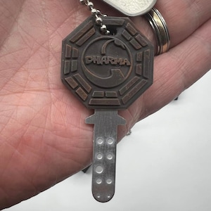 May include: A brown metal key chain with a Dharma Initiative logo and a silver key. The key has a silver handle and a silver blade with six holes.