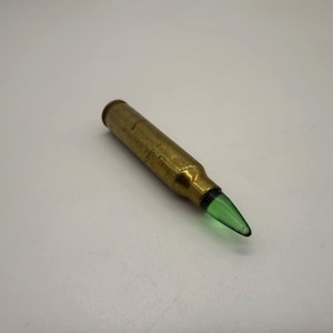 May include: A brass bullet-shaped bottle stopper with a green glass tip. The stopper is made of brass and has a metallic finish. The green glass tip adds a pop of color to the design. The bottle stopper is designed to fit standard wine bottles.