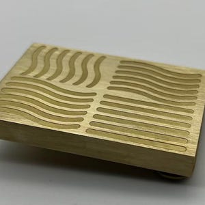 Könnte beinhalten: Rechteckiger Gegenstand aus messingfarbenem Metall mit geätzten geschwungenen und geraden Linien. Das Design ist symmetrisch, mit wellenartigen Mustern. Das Objekt scheint ein dekoratives Element oder ein kleiner, fester Block zu sein.