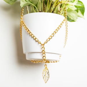 Peut inclure: Un pot de fleurs blanc suspendu avec une chaîne dorée et un pendentif en forme de feuille. La chaîne est attachée au pot et pend vers le bas, avec le pendentif en forme de feuille en bas.