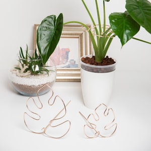 Pode incluir: Dois suportes de plantas de arame dourado em forma de folhas de monstera. Os suportes estão sobre uma superfície branca. Uma planta em vaso com folhas verdes está no fundo.