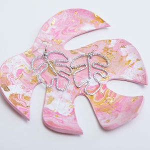 May include: A pair of silver beaded earrings shaped like monstera leaves, displayed on a pink and gold leaf-shaped dish.