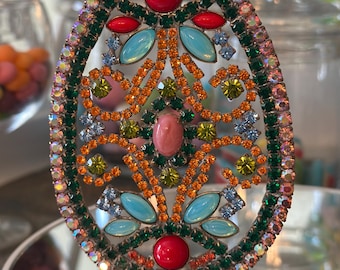 May include: A colorful jeweled Easter egg ornament with a green, orange, and blue design. The egg is on a gold stand and has a red, pink, and blue jeweled design.
