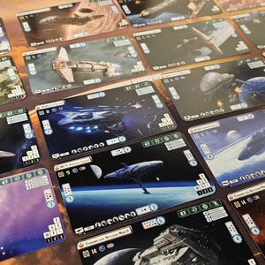 Può includere: Una collezione di carte da gioco rettangolari con illustrazioni di astronavi. Le carte hanno uno sfondo scuro con immagini di astronavi, pianeti e battaglie spaziali. Ogni carta include testo e simboli.
