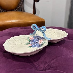 May include: Two off-white, oval-shaped ceramic dishes with a floral design and a blue ribbon handle. The dishes are placed on a purple velvet ottoman, with a brown leather chair in the background.
