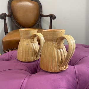 May include: Two ceramic mugs with brown and beige speckled glaze. The mugs have a ribbed design and a handle with a star-shaped design.
