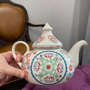 May include: White ceramic teapot with a lid and a handle. The teapot is decorated with a colorful floral pattern in shades of red, blue, green, and purple.