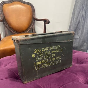 May include: An olive green metal ammunition box with the text "200 CARTRIDGES 7.62 MM, M13 CARTONS, 1 M62-4 M80 LC-92M602L011". The box is rectangular with a hinged lid and is sitting on a purple tufted ottoman.